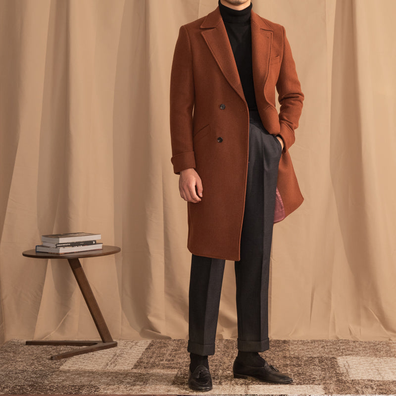 Person wearing a brown coat standing in front of a beige curtain with a small table and books in the background.
